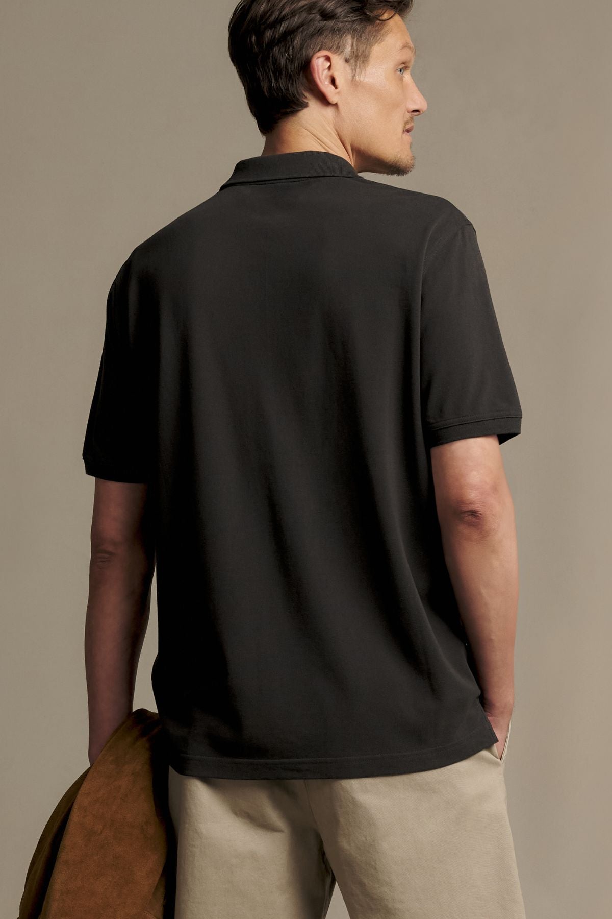A man wearing the RM Williams Rod Polo in black and beige pants stands facing away, holding a brown jacket in his left hand.