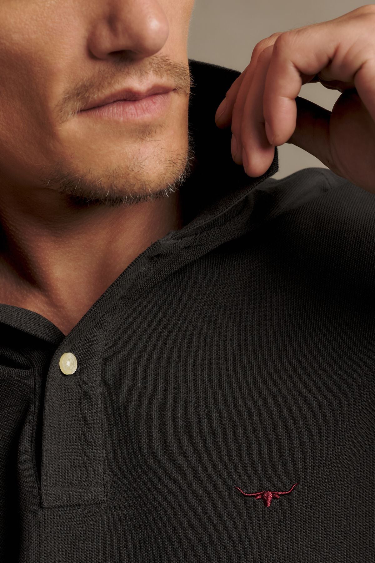 A close-up of a man wearing the RM Williams Rod Polo, featuring a black collared design with a small red bull logo embroidered on the chest. He is adjusting the collar with his hand and has light facial hair.
