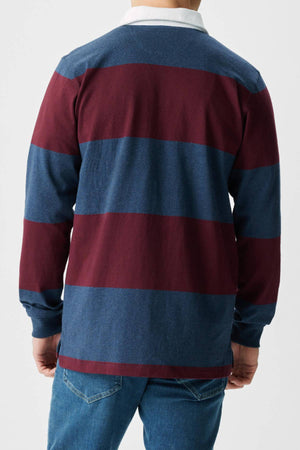 Someone is shown from the back against a plain light background, wearing blue jeans and the RM Williams Tweedale Rugby—a long-sleeve polo in organic cotton with wide maroon and navy blue stripes.