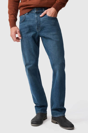 A person stands against a plain light background wearing a brown sweater, black shoes, and Rodd & Gunn Kurow Relaxed Fit Italian Denim Jeans in blue, with one hand in their pocket. Their face is not visible.
