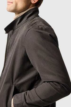 A man in a Rodd & Gunn Armitage Jacket with YKK zips over a light turtleneck stands sideways with one hand in his pocket against a plain light background.