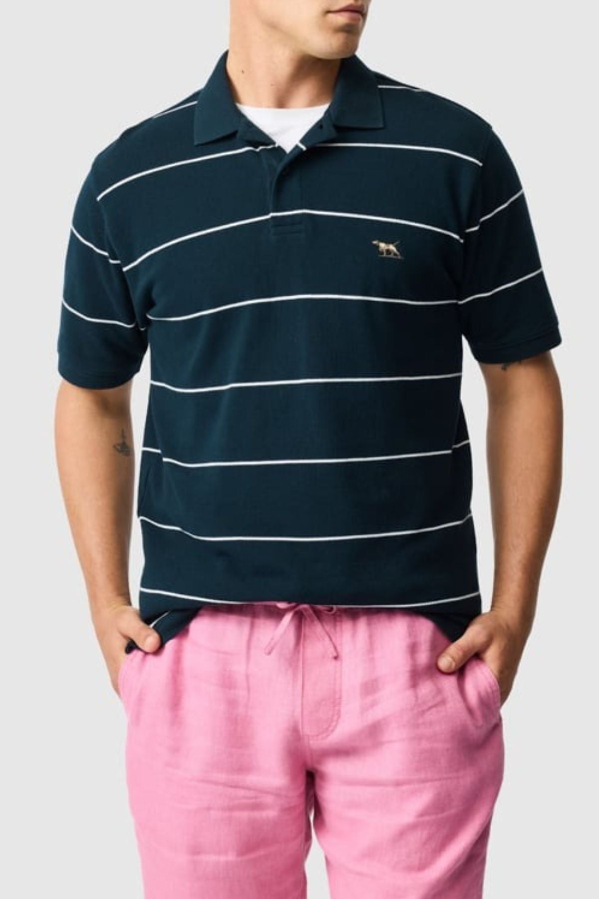 A person wearing a Rodd & Gunn Glen Eden Polo with white horizontal stripes and pink pants stands against a plain background, hands tucked into pockets. The navy blue shirt features a high-retention rib collar for added style and comfort.