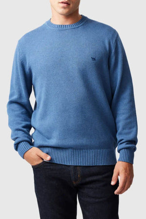 A man stands with one hand in his pocket against a plain light background, wearing the Rodd & Gunn Gunn Knit—a stylish, comfortable 100% cotton light blue crew neck sweater with long sleeves—and dark blue jeans.
