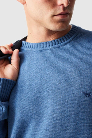 A close-up shows a man wearing the Rodd & Gunn Gunn Knit, a stylish and comfortable blue crew neck sweater, with a black strap over his shoulder; only his lower face and upper torso are visible.