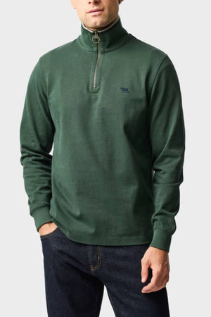 A man wears a dark green Rodd & Gunn Gunn 14oz 1/4 Zip Sweat made from 100% Australian Good Earth Cotton with FibreTrace® technology, paired with dark blue jeans, standing with one hand in his pocket against a plain light background.