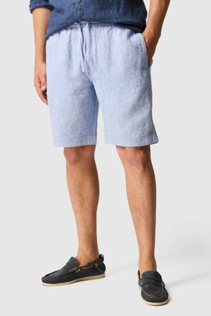 A person stands with hands in pockets, wearing light blue Rodd & Gunn Linen Resort Shorts, a blue shirt with rolled sleeves, and black loafers against a plain light gray background.