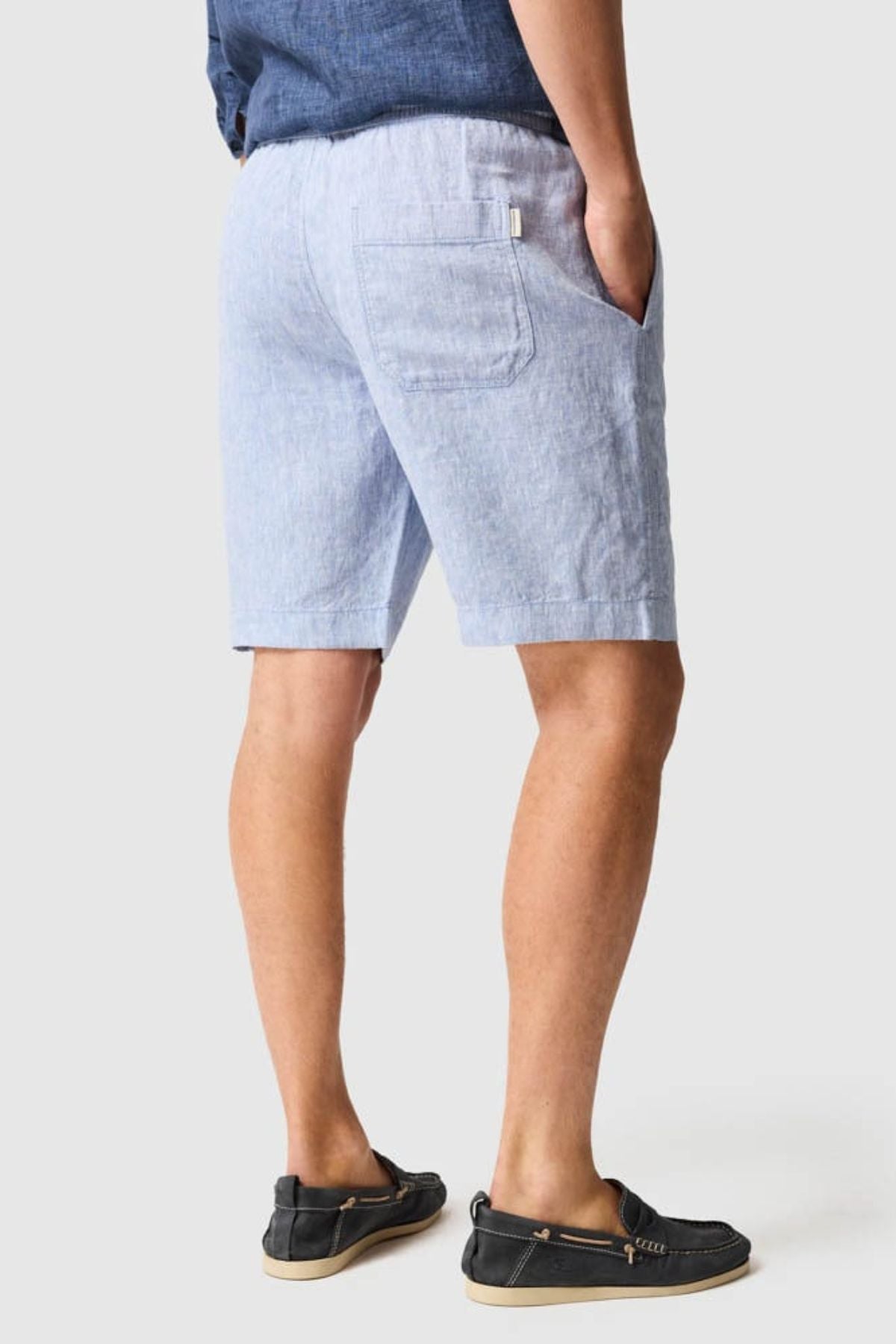 A man faces away from the camera, wearing Rodd & Gunn Linen Resort Shorts in light blue, a blue shirt, and black boat shoes with beige soles. His right hand is in his pocket against a plain light grey background.