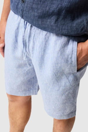 Close-up of a person wearing Rodd & Gunn Linen Resort Short in light blue with hands in pockets, paired with a dark blue shirt. Only the lower torso and upper legs are visible against a plain background.