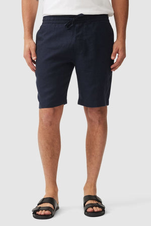 A person wearing a white shirt, Rodd & Gunn Linen Resort Short in dark navy, and black sandals stands against a plain light background with their hands relaxed by their sides.