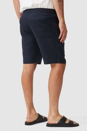 A person wearing a white shirt, Rodd & Gunn Linen Resort Short in navy blue, and black sandals is shown from the back with one hand by their side. A small tattoo appears on their upper left arm.