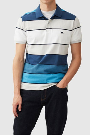 A person dons a Rodd & Gunn McLeod Bay Sports Fit Polo in blue and white stripes, highlighting the cotton polo's pique texture. They pair it with dark jeans, resting their left hand in their pocket against a plain gray background.