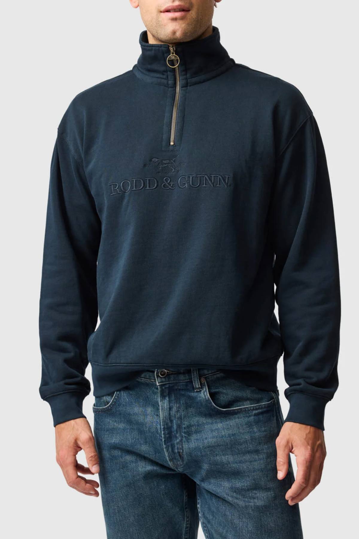 A man wears the Rodd & Gunn Milford Sound Sweat in navy blue, with "RODD & GUNN" embroidered on the chest, a YKK metal zip, and blue jeans, standing against a plain light gray background.