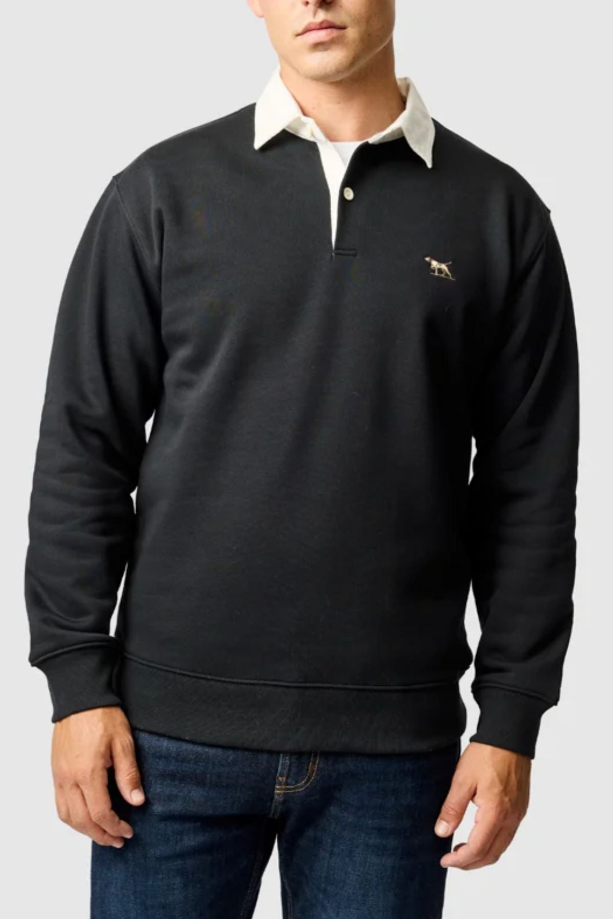 A man wearing a dark Rodd & Gunn Otago Sweat with a classic twill rugby collar and signature chest logo, paired with blue jeans, stands against a plain light gray background.