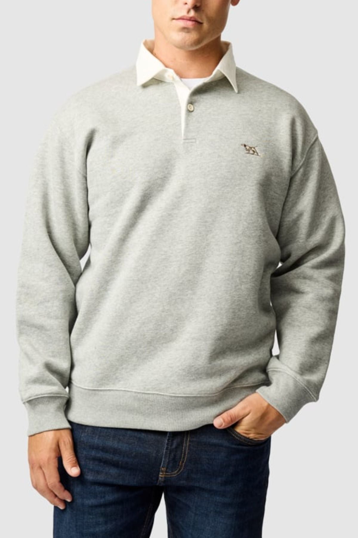 A man in a Rodd & Gunn Otago Sweat, styled with a white collared shirt and dark blue jeans, showcases the signature pointer logo while standing against a plain light gray background.