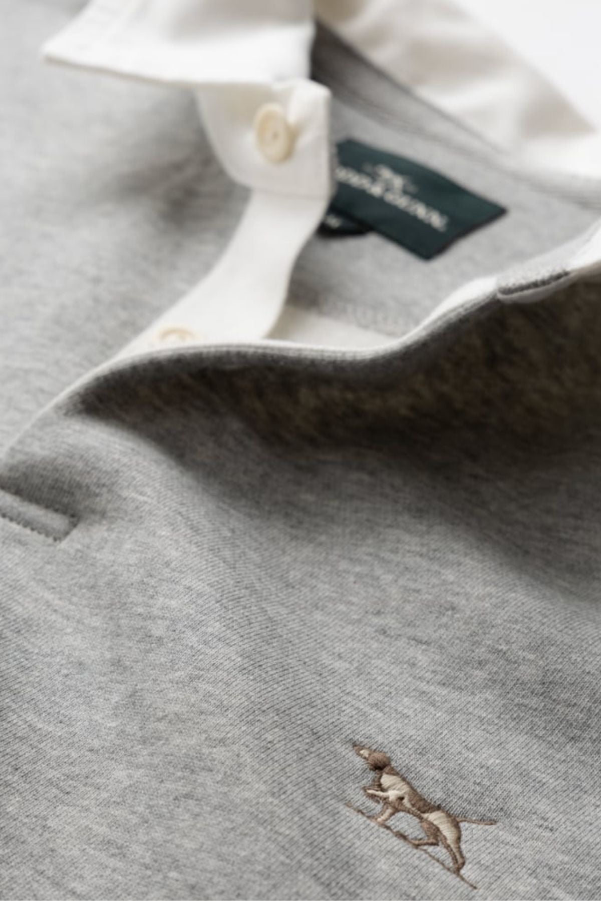 Close-up of the Rodd & Gunn Otago Sweat in light grey, featuring a traditional twill rugby collar with buttons, signature pointer logo embroidered on the chest, and a dark green tag inside the collar.