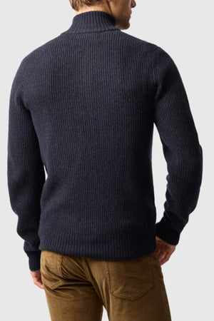 A man stands facing away, wearing the Rodd & Gunn Robbies Road Knit sweater in dark blue with brown pants. The background is plain and light-colored.