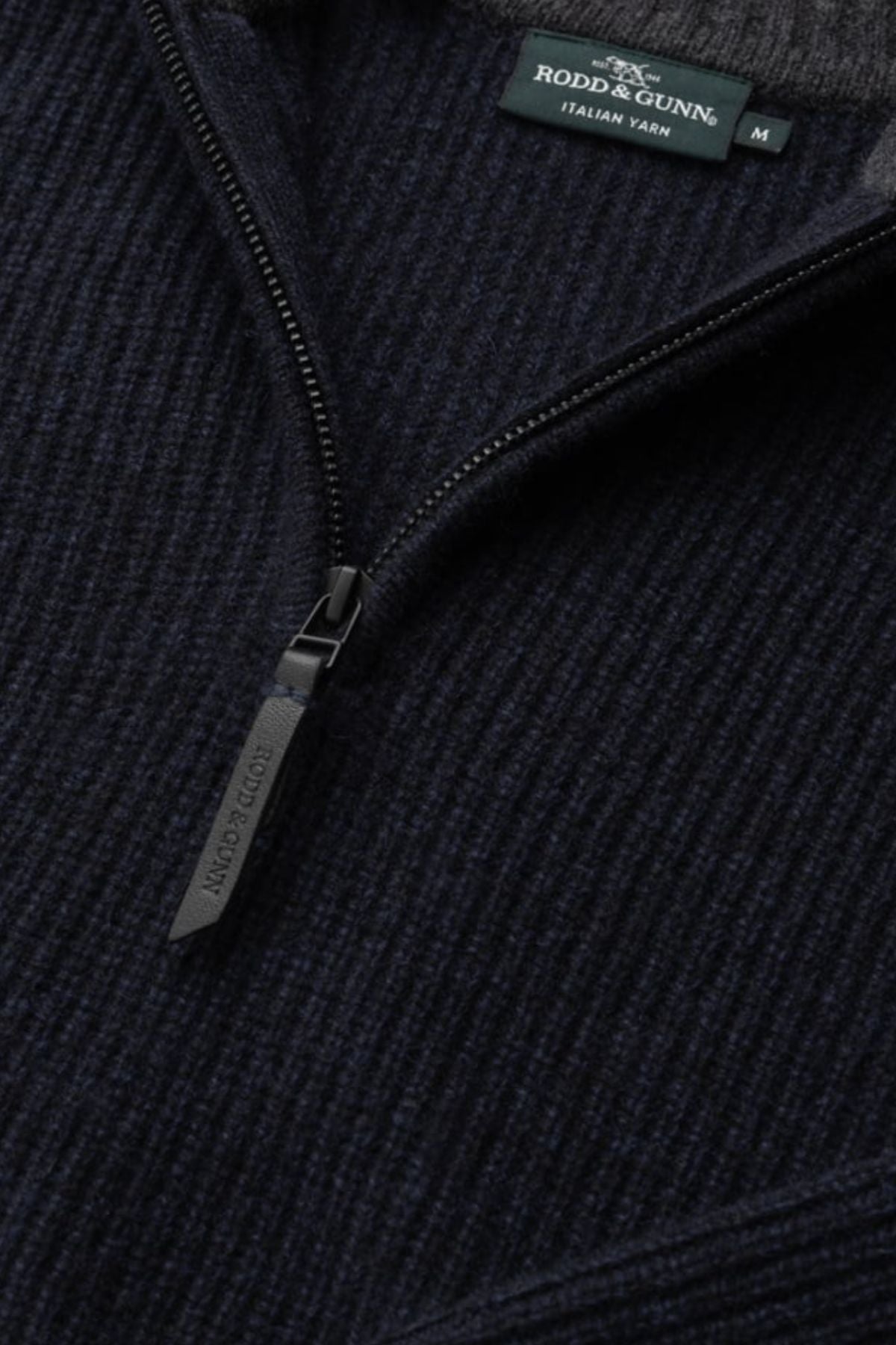 Close-up of the Rodd & Gunn Robbies Road Knit, a dark navy quarter-zip sweater in fisherman's rib knit, featuring a visible zipper pull and tag. Made from premium New Zealand Merino Wool.