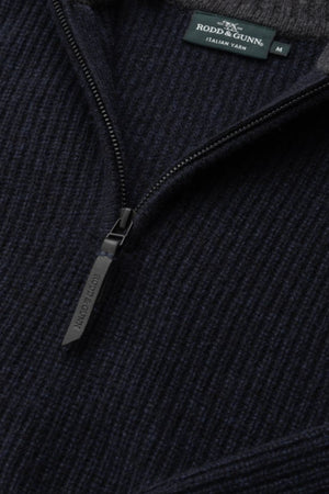 Close-up of the Rodd & Gunn Robbies Road Knit, a dark navy quarter-zip sweater in fisherman's rib knit, featuring a visible zipper pull and tag. Made from premium New Zealand Merino Wool.