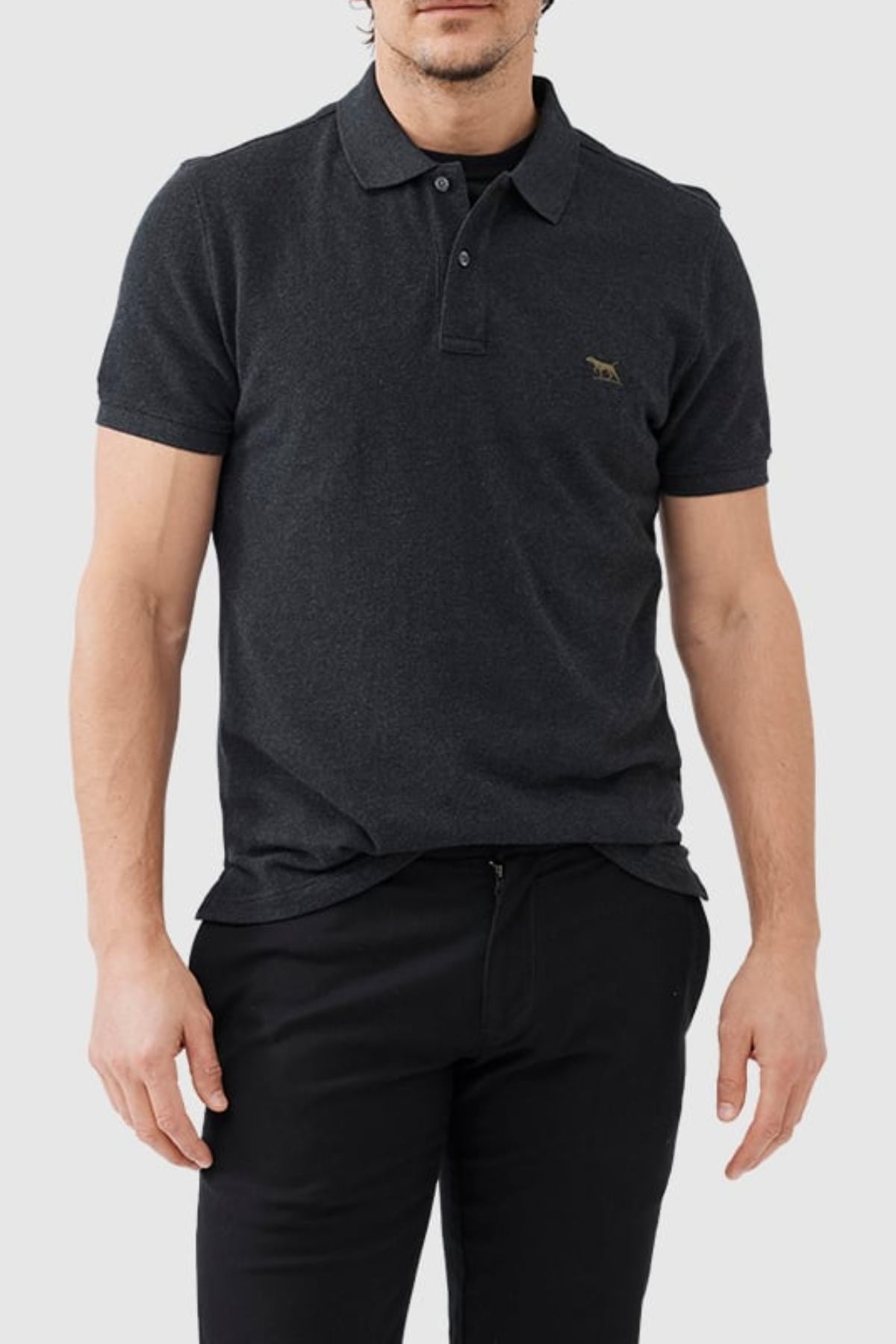 A man wearing the Rodd & Gunn The Gunn Polo in dark grey with a small embroidered animal logo on the chest, paired with black pants, stands against a light grey background.