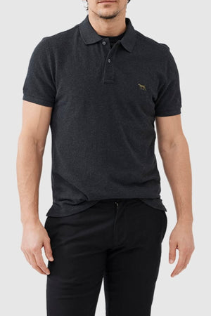 A man wearing the Rodd & Gunn The Gunn Polo in dark grey with a small embroidered animal logo on the chest, paired with black pants, stands against a light grey background.