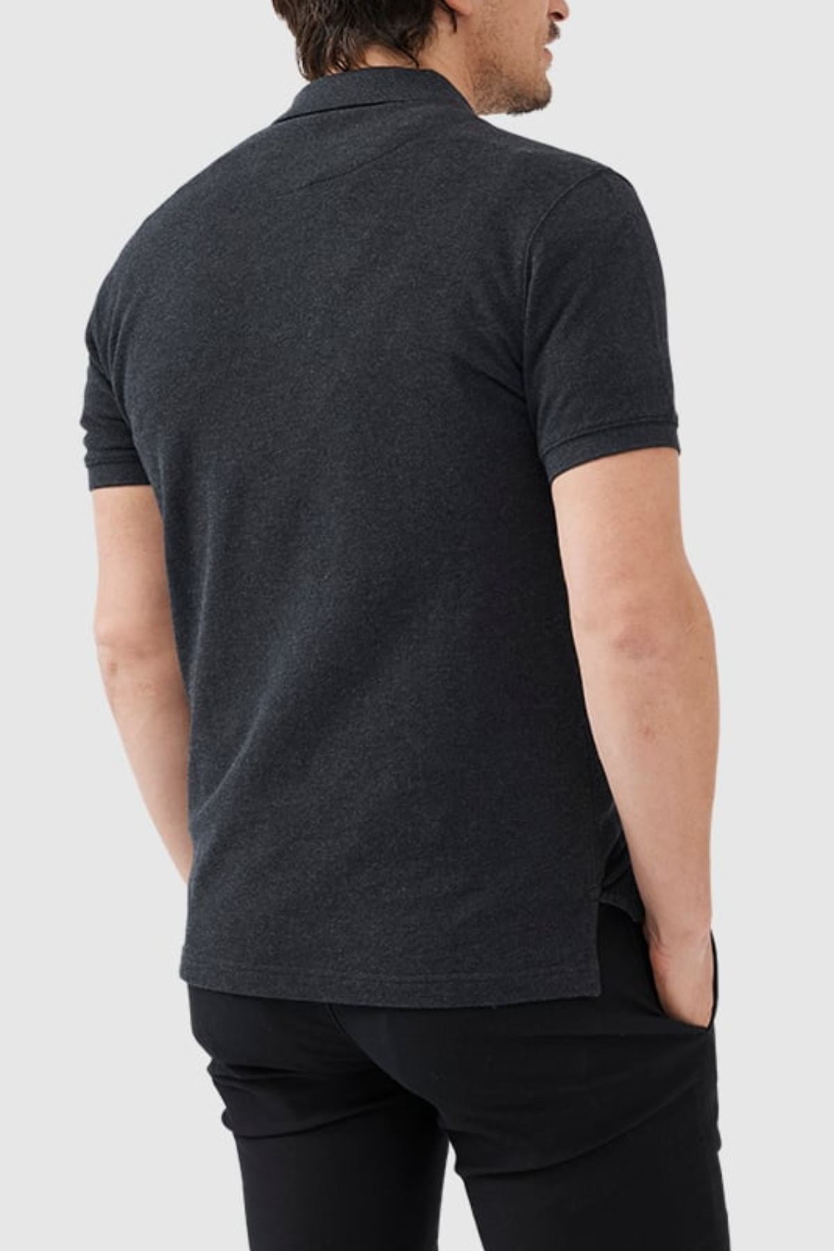 A man stands with his hands in his pockets, facing away from the camera, wearing a Rodd & Gunn The Gunn Polo in dark gray and black pants against a light gray background.