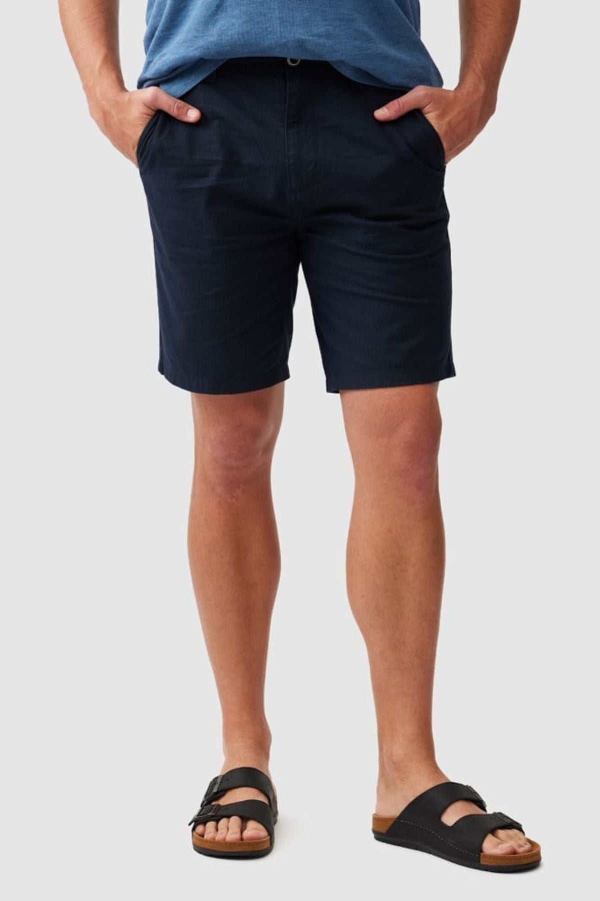 A person stands with hands in pockets against a plain light gray background, wearing a classic fit blue shirt, black sandals, and Rodd & Gunn The Gunn Short in dark navy.