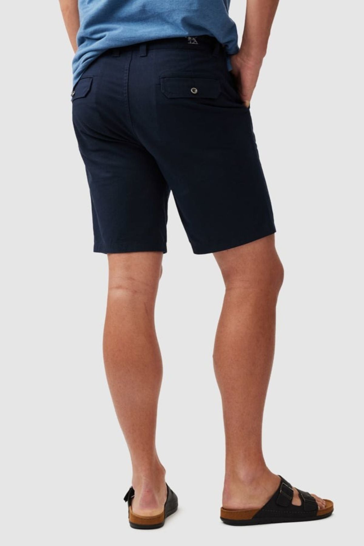 Wearing a blue classic fit shirt, Rodd & Gunn The Gunn Short in dark navy, and black sandals, a person stands with their back to the camera against a light gray background. The shirt is made from soft cotton elastane for comfort and movement.