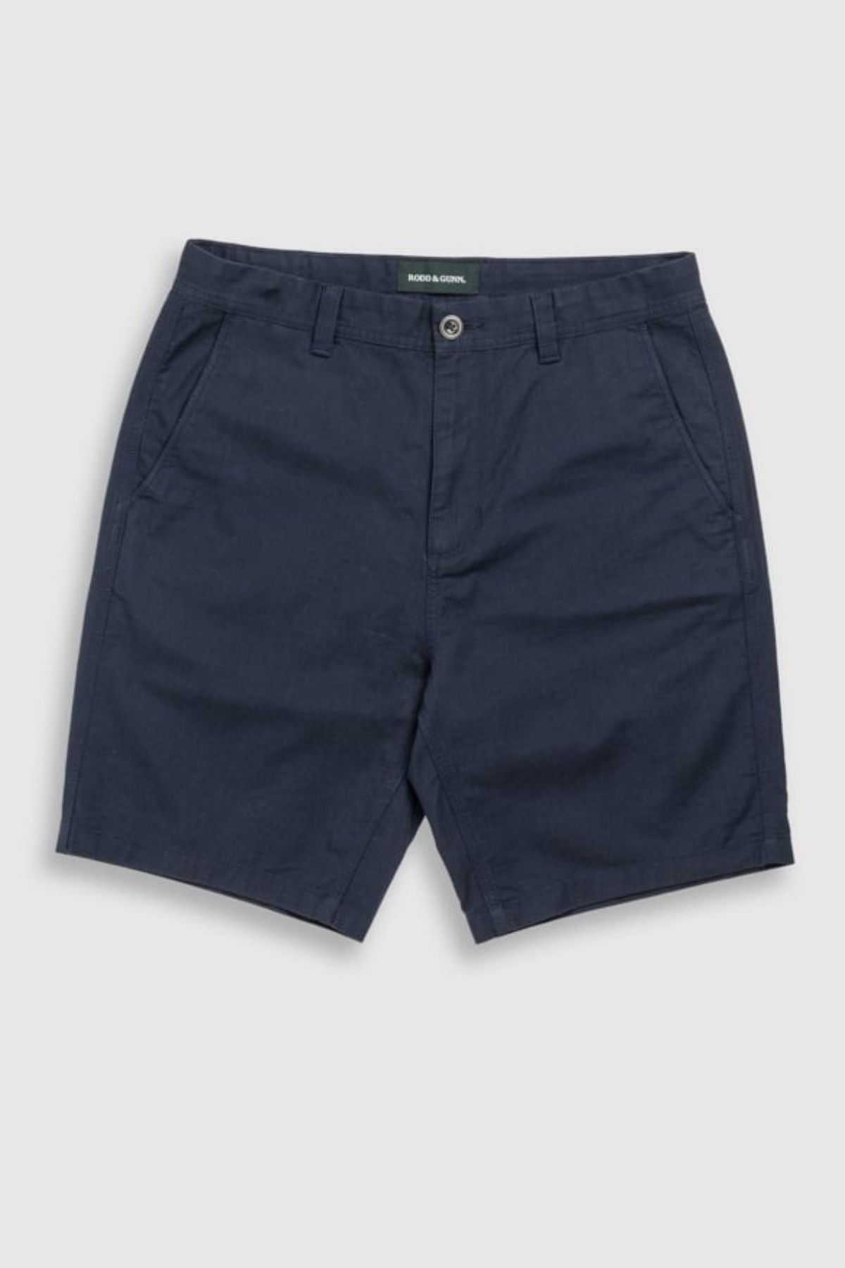 The Rodd & Gunn The Gunn Short is a pair of navy blue classic fit chino shorts with side pockets, button and zip closure, belt loops, and subtle logo detailing, shown on a light grey background.