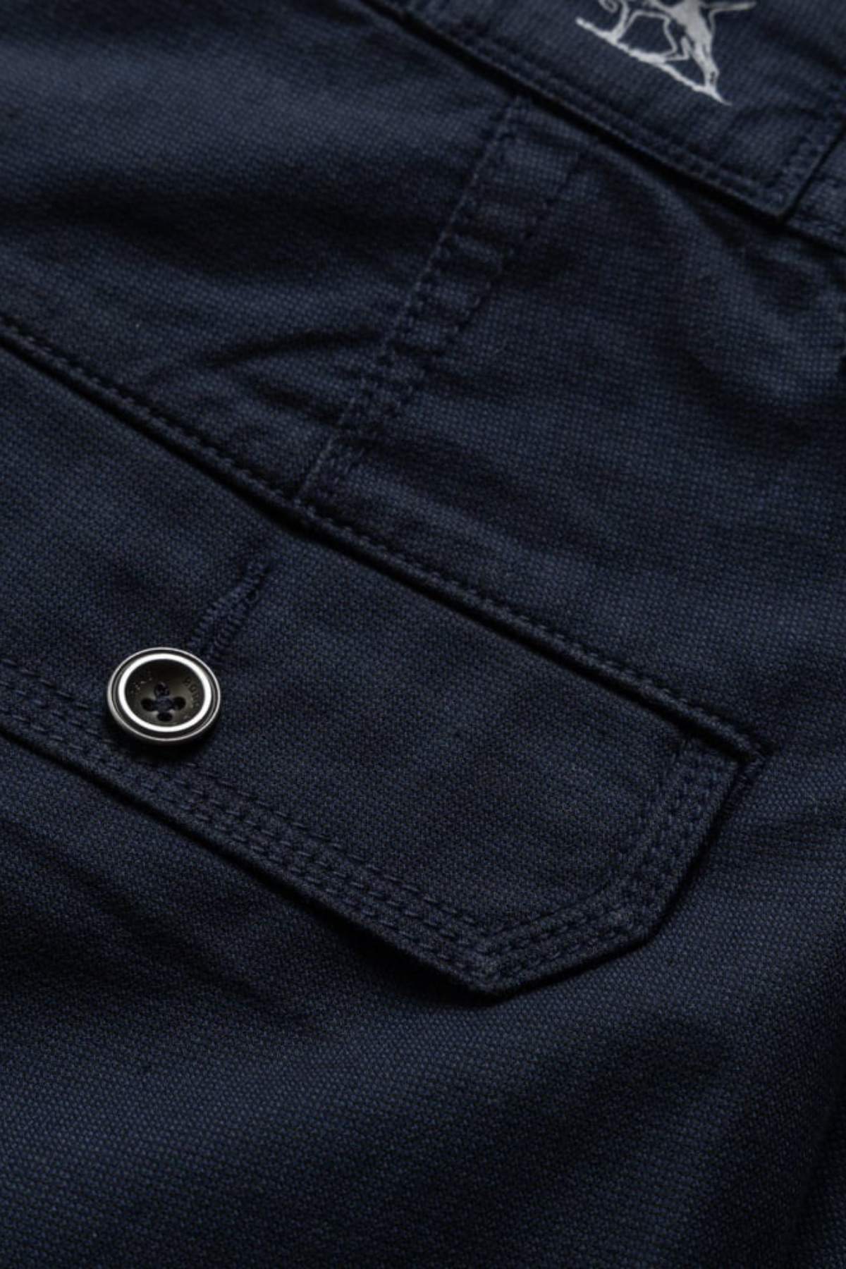 Close-up of a dark navy blue pocket with flap, black button, and visible stitching on the Rodd & Gunn The Gunn Short. Made from cotton elastane in a classic fit, it features part of the white Rodd & Gunn logo embroidered near the top.