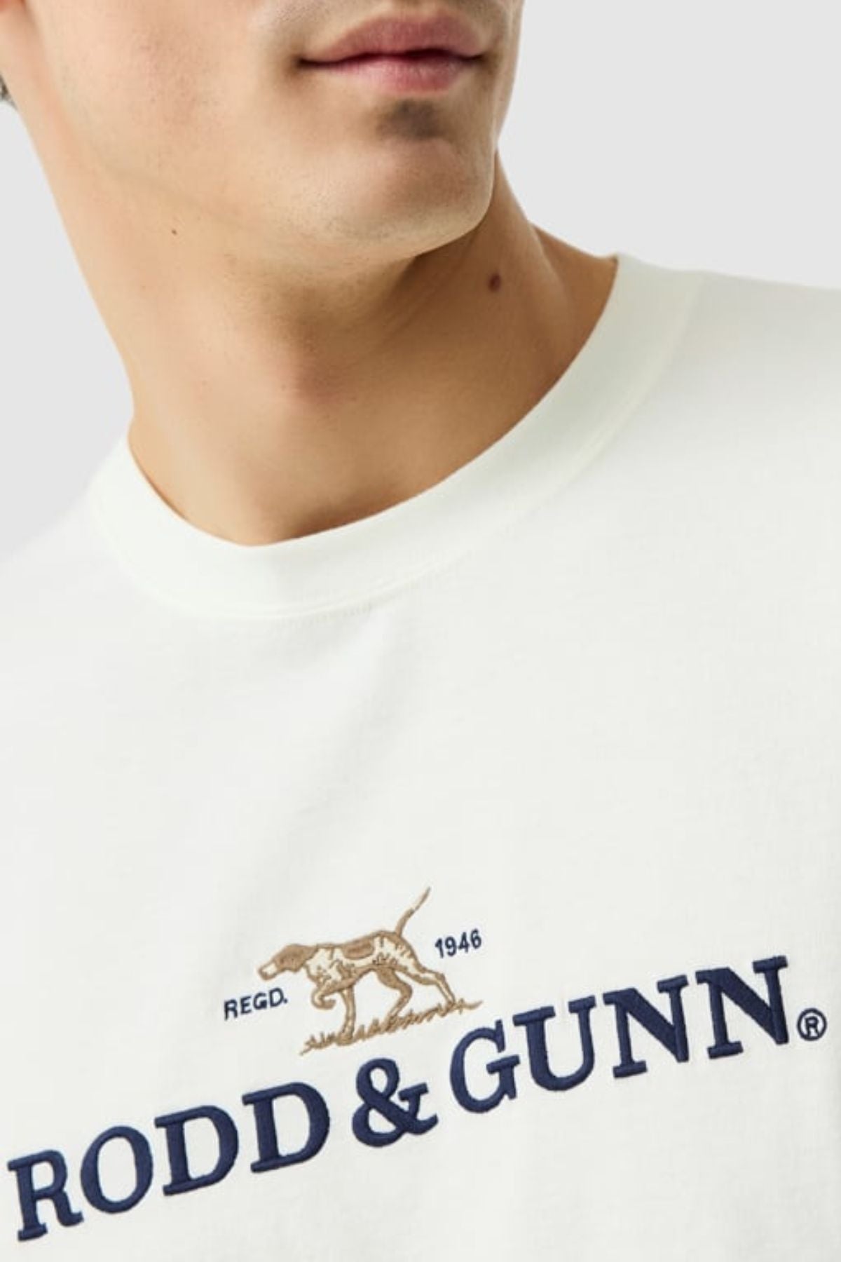 A person wears the Rodd & Gunn The Peaks Logo Tee, a classic white T-shirt with a brown dog logo, "1946," and bold navy "RODD & GUNN" text. The photo shows the upper torso and highlights this FibreTrace technology shirt.