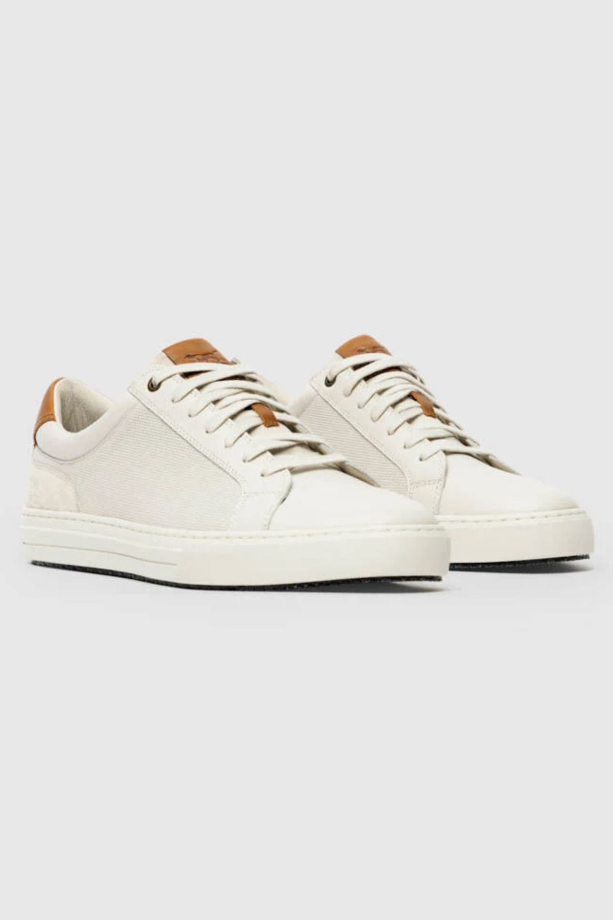 The Rodd & Gunn Transit Sneaker is an off-white low-top shoe with white soles and laces, featuring brown leather accents on the tongue and heel plus a removable foot bed, shown on a light gray background.