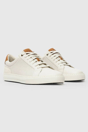 The Rodd & Gunn Transit Sneaker is an off-white low-top shoe with white soles and laces, featuring brown leather accents on the tongue and heel plus a removable foot bed, shown on a light gray background.