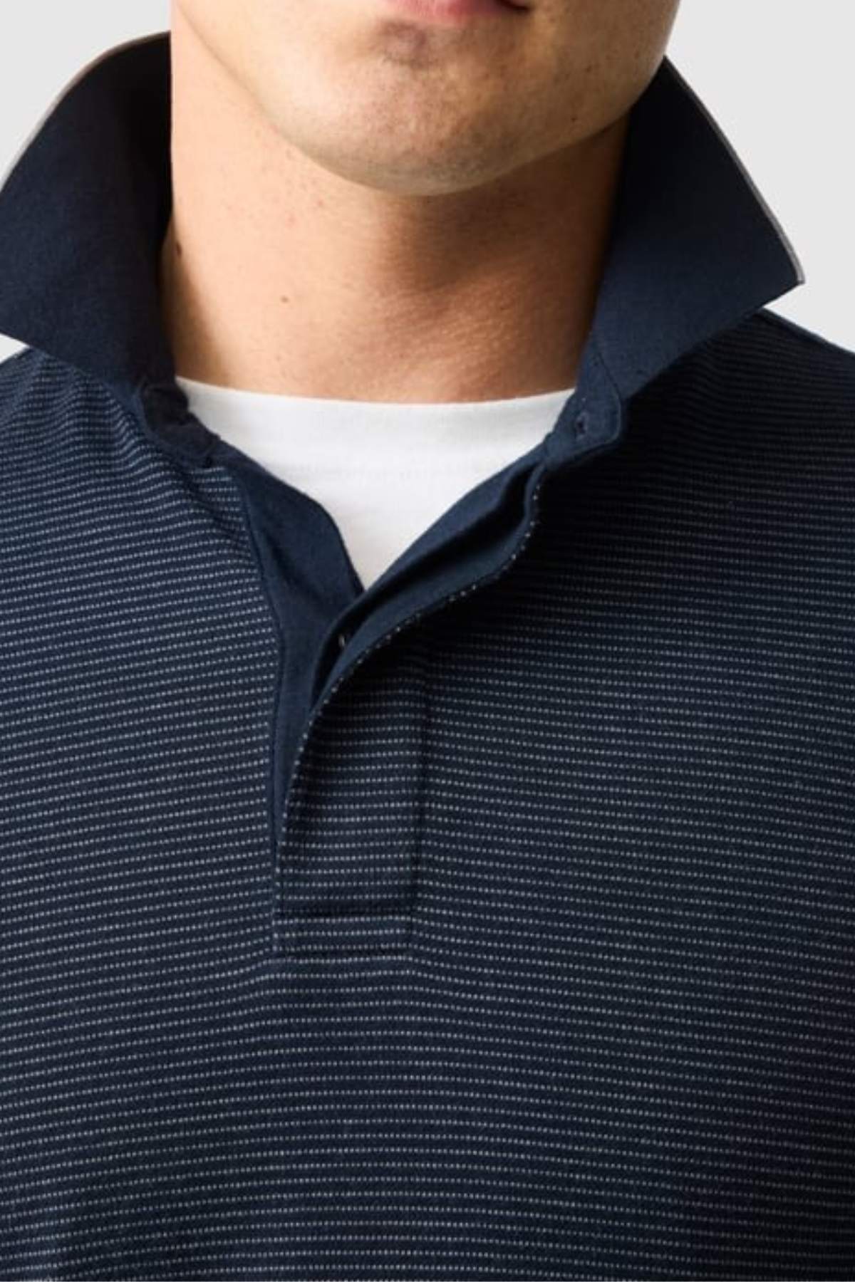 A close-up shows a person in the Rodd & Gunn Wilson Bay Sports Fit Polo, a breathable birdseye knit shirt with dark pinstripes and a popped collar over a white crew-neck tee. Only their lower face and upper torso are visible.