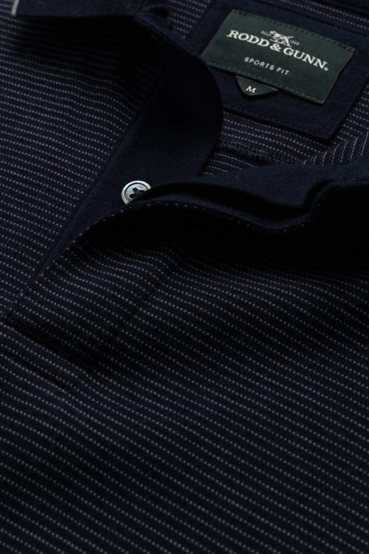 Close-up of the Rodd & Gunn Wilson Bay Sports Fit Polo in dark navy with thin white stripes and a visible "Sports Fit" label near the collar. Part of the button placket is also featured.