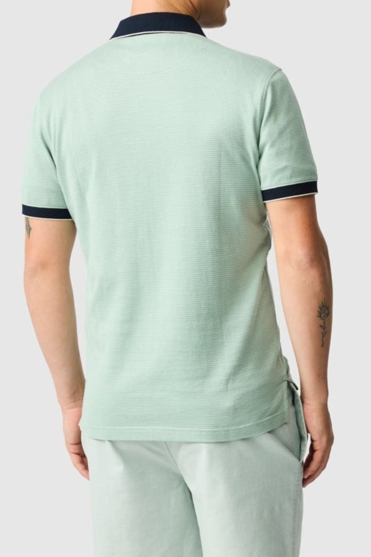 A person is wearing a light green Rodd & Gunn Wilson Bay Sports Fit Polo with dark collar and sleeve trim, paired with matching shorts. Small tattoos decorate their forearm and bicep as they stand against a plain light gray background, facing away.