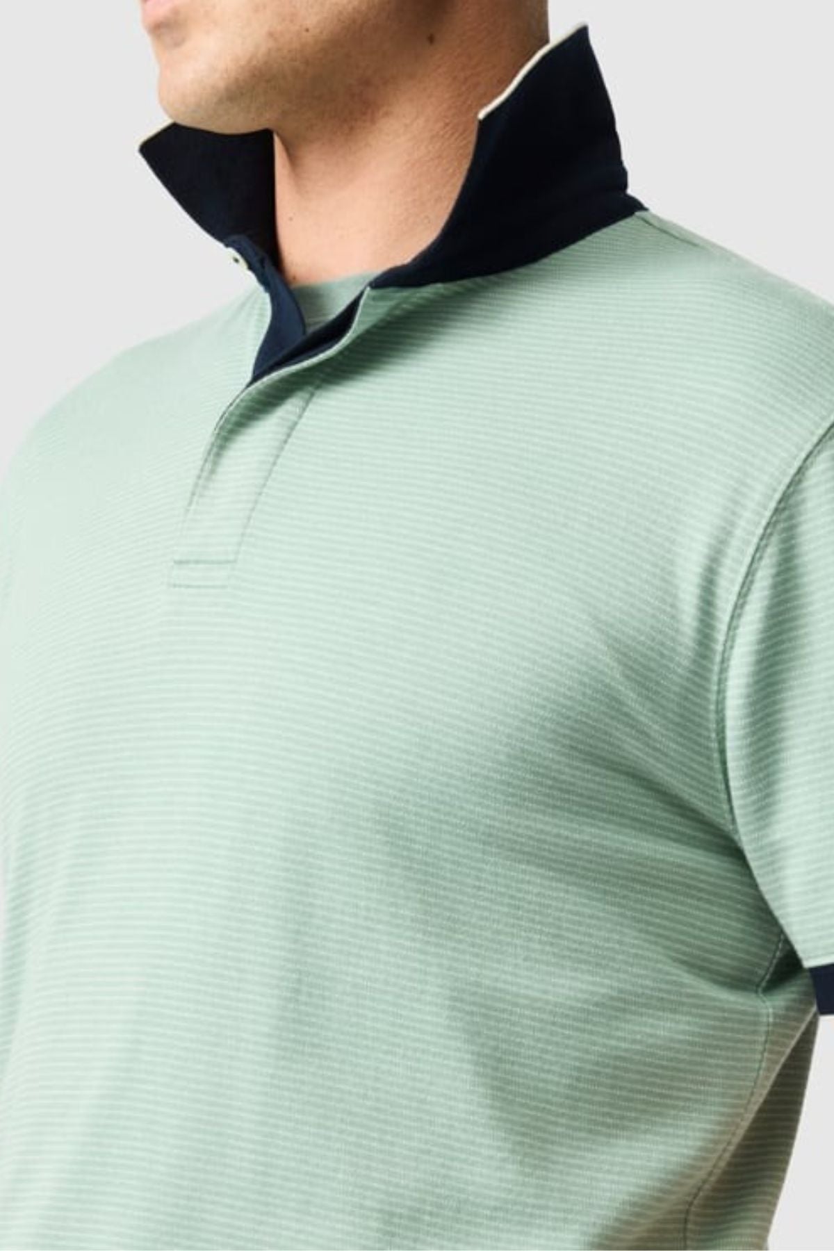 A person is shown from shoulders to just above the midriff, facing slightly right, wearing a light green Rodd & Gunn Wilson Bay Sports Fit Polo featuring a breathable birdseye knit with a dark blue inner collar. The plain gray background accentuates the polo's sleek design.