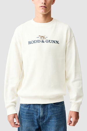 Someone wears the Rodd & Gunn Bromley Sweat, a sustainable sweatshirt made with recycled polyester. It has long sleeves, a light gray color, and features the brand name plus a small dog logo on the chest, paired with blue jeans.