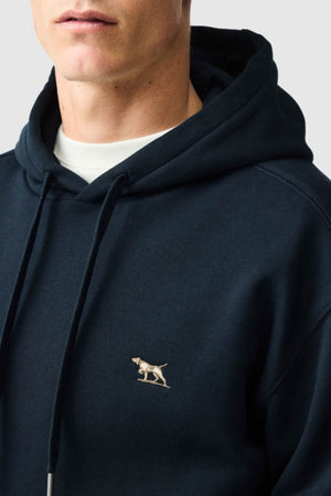 A person is wearing a dark blue Rodd & Gunn Otago Hoodie, featuring a small embroidered dog logo on the chest. Crafted from soft Australian Cotton with cozy brushed back fleece, the hoodie includes drawstrings. The person is positioned slightly sideways against a plain light gray background.