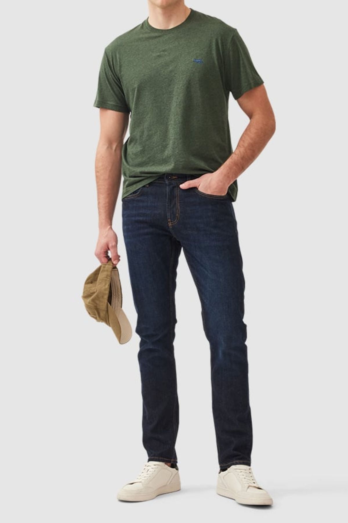 A person wearing the Rodd & Gunn The Gunn T-Shirt, dark jeans, and white sneakers holds a beige cap. They're in a sports fit green T-shirt with one hand in their pocket, standing against a plain white background that highlights their relaxed style crafted from Australian Cotton.