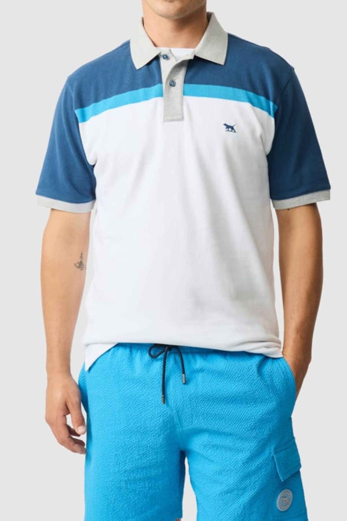 A person wears the Rodd & Gunn Whitby Polo with a color block design in white, blue, and gray, paired with bright blue drawstring shorts. They pose with one hand in their pocket against a plain light background.