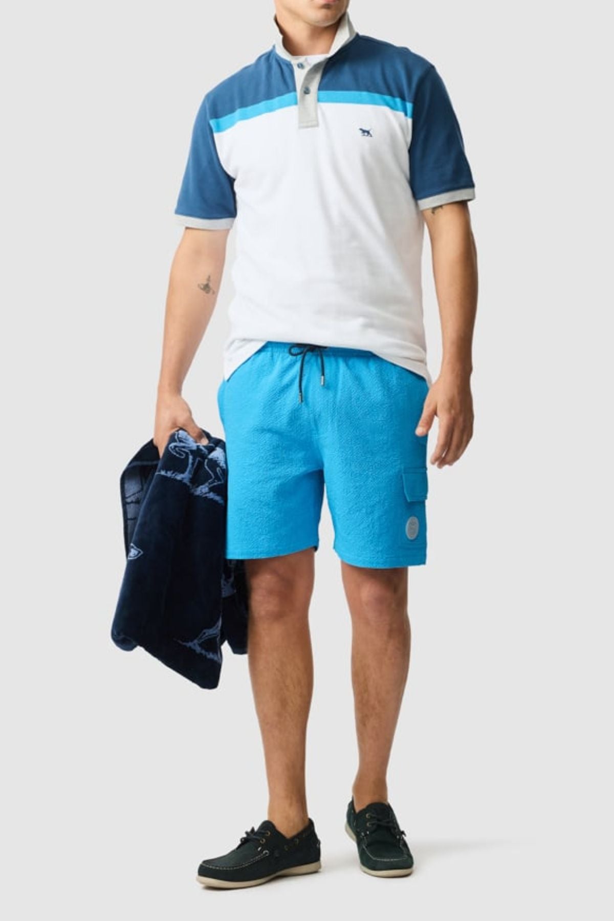 A man wears a blue and white Rodd & Gunn Whitby Polo with color block design, bright blue shorts, and black boat shoes while holding a navy jacket against a plain light gray background.