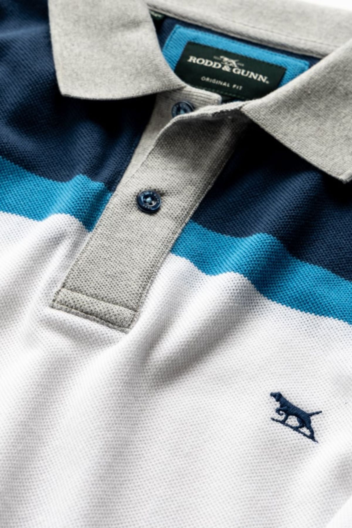 Close-up of the Rodd & Gunn Whitby Polo, featuring grey, navy blue, teal, and white horizontal color block stripes, blue buttons, and an embroidered dog logo near the hem. The label reads "Rodd & Gunn.