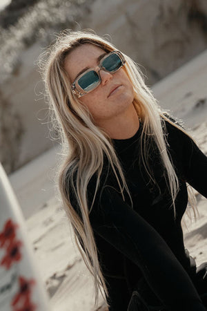 A woman with long blonde hair wears SafeStyle Auras Graphite Frame Grey Polarised Lens sunglasses and a black wetsuit while sitting on a sandy beach, gazing slightly upward. The background features blurred sand and dunes.