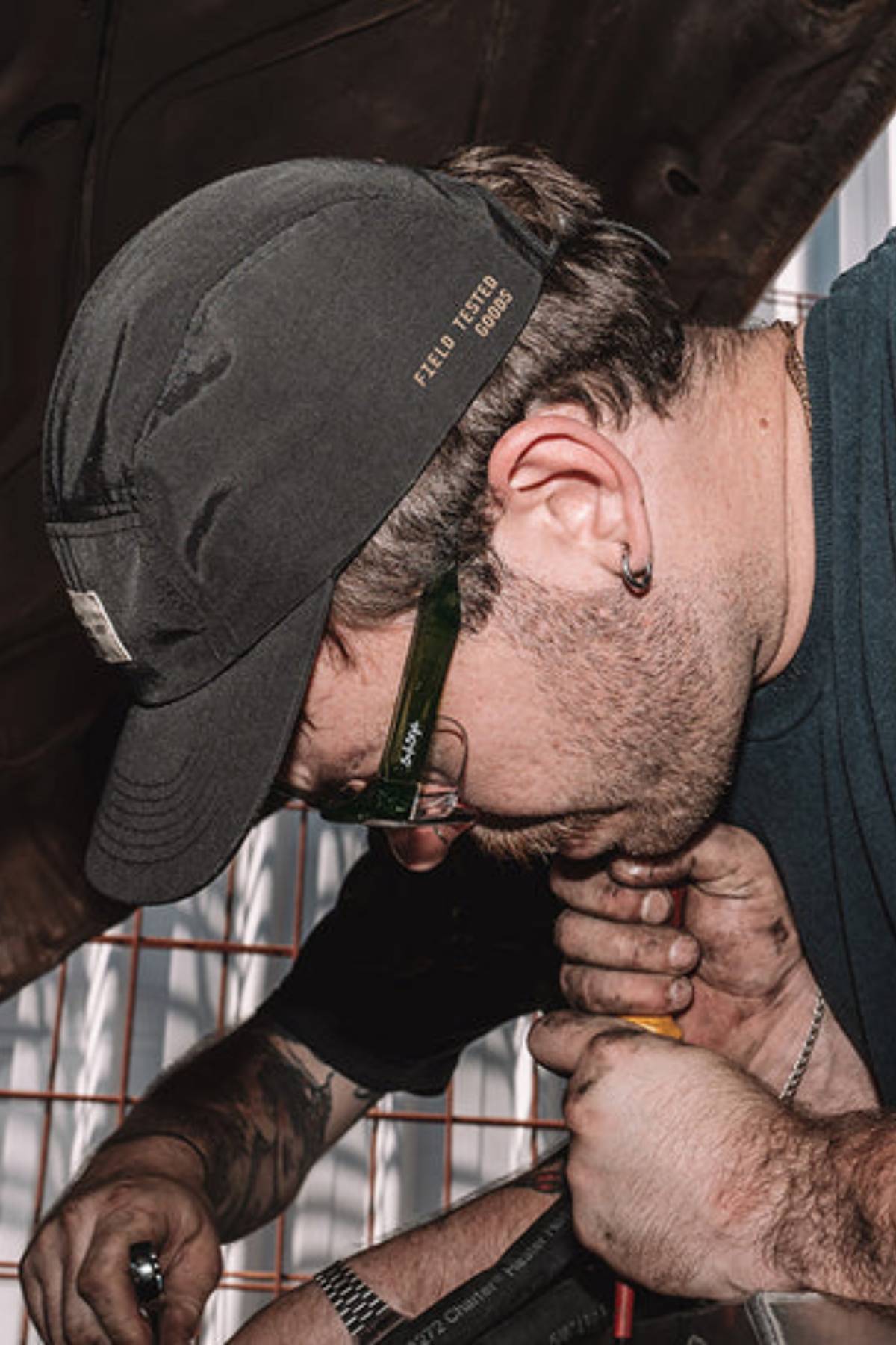 A man wearing SafeStyle Classics Green Frame Polarised Lens glasses, a black cap, and a dark shirt is intensely focused on a mechanical task, using both hands on a tool while another tattooed arm assists him.