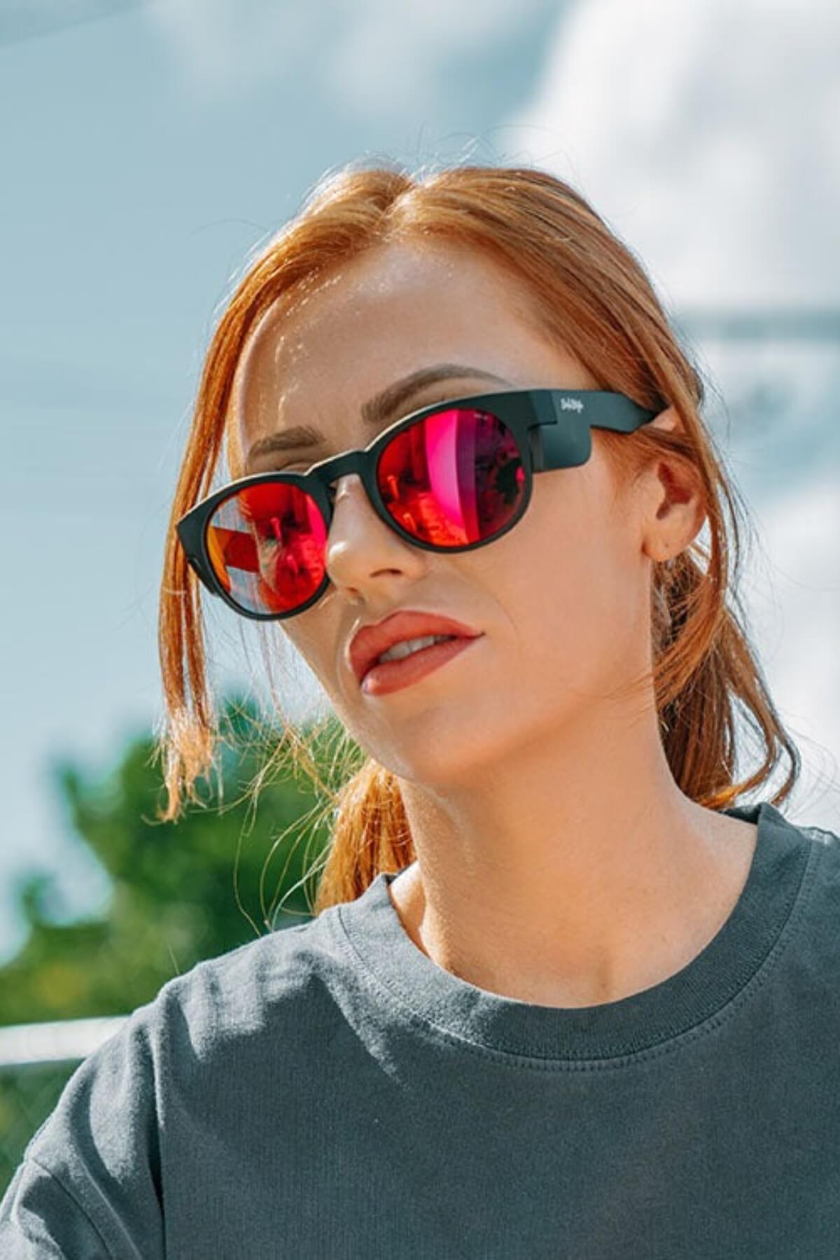 A red-haired woman sports SafeStyle Cruisers with a matte black frame and mirror red polarized lenses offering UV protection, wearing a grey T-shirt. She stands outdoors beneath a blue sky dotted with fluffy clouds.