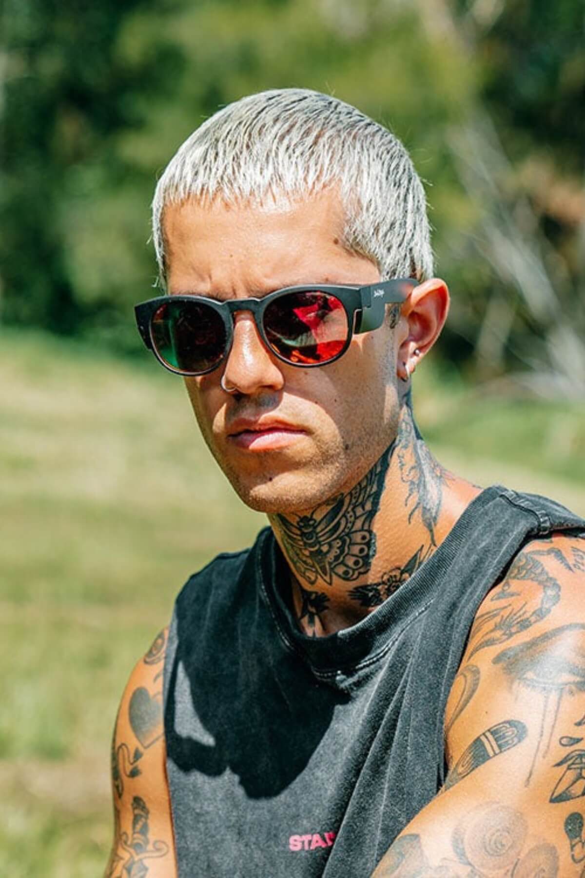 A person with short, bleached hair and SafeStyle Cruisers Matte Black Frame Mirror Red Polarised Lens sunglasses enjoys the outdoors. They're wearing a sleeveless black shirt, revealing tattoos on their arms and neck against blurred greenery that provides natural UV protection on a sunny day.
