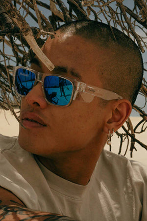 A person with a shaved head and facial hair wears SafeStyle Fusions Clear Frame Mirror Blue Polarised Lens sunglasses, relaxing under tree branches on a sandy beach. The stylish glasses reflect the coast and offer UV protection with clear, polarised vision.