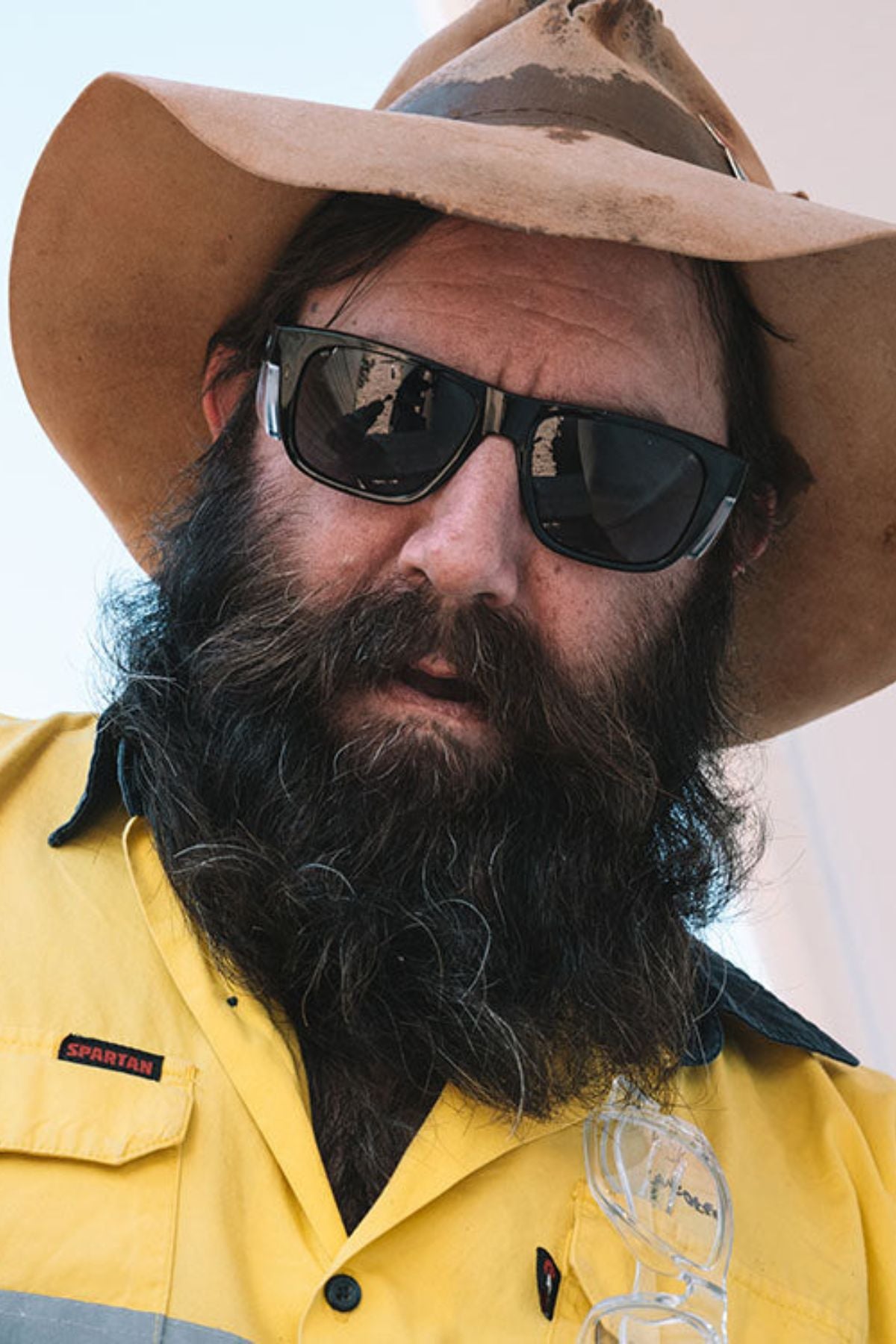A man with a long beard in a yellow shirt and cowboy hat is wearing dark sunglasses, specifically SafeStyle Fusions XL Black Frame Polarised Lens, which offer UV protection.
