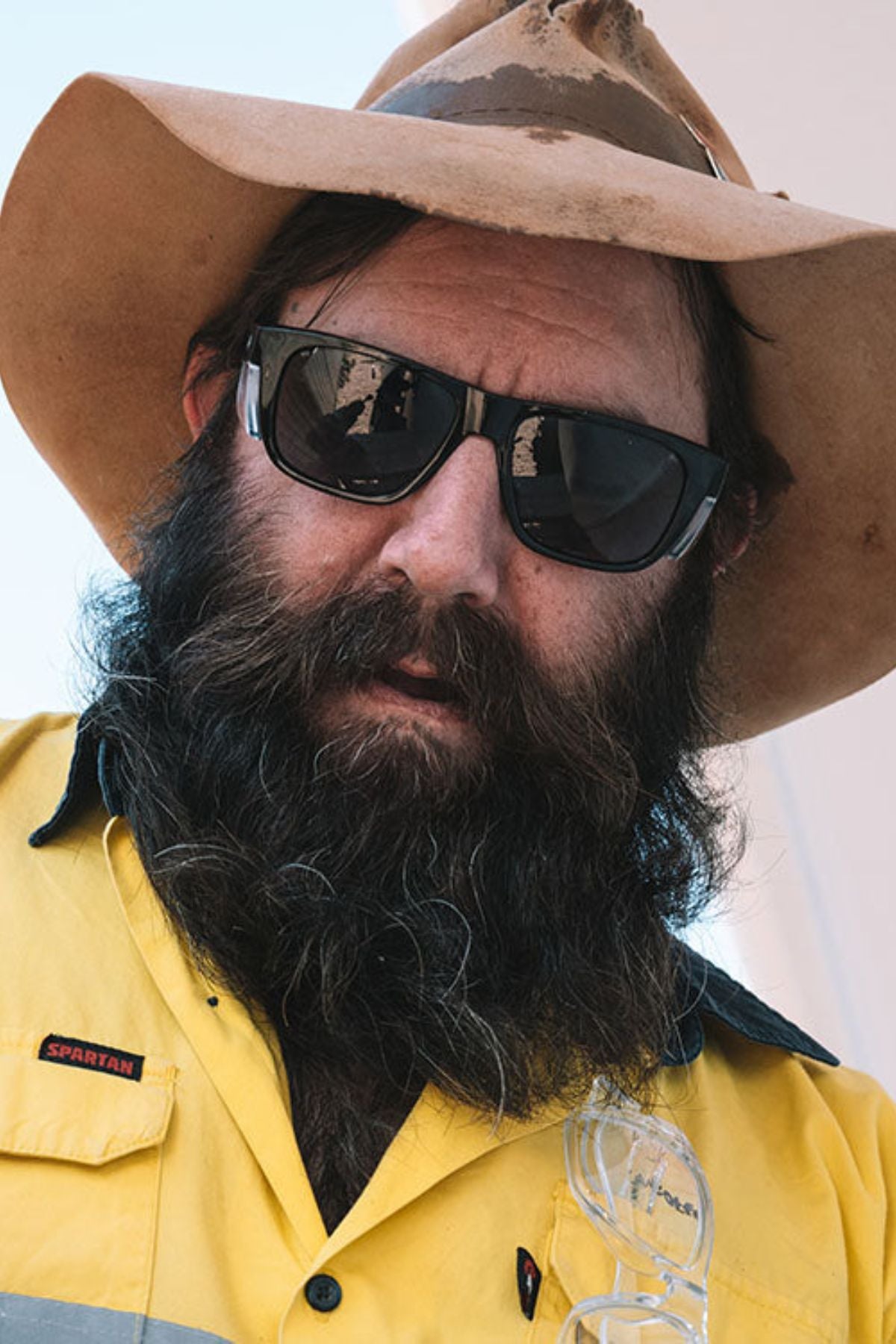 A man with a thick beard wears a wide-brimmed hat, SafeStyle Fusions XL Black Frame Tinted Lens sunglasses, and a bright yellow "Spartan" work shirt; safety glasses hang from his collar.