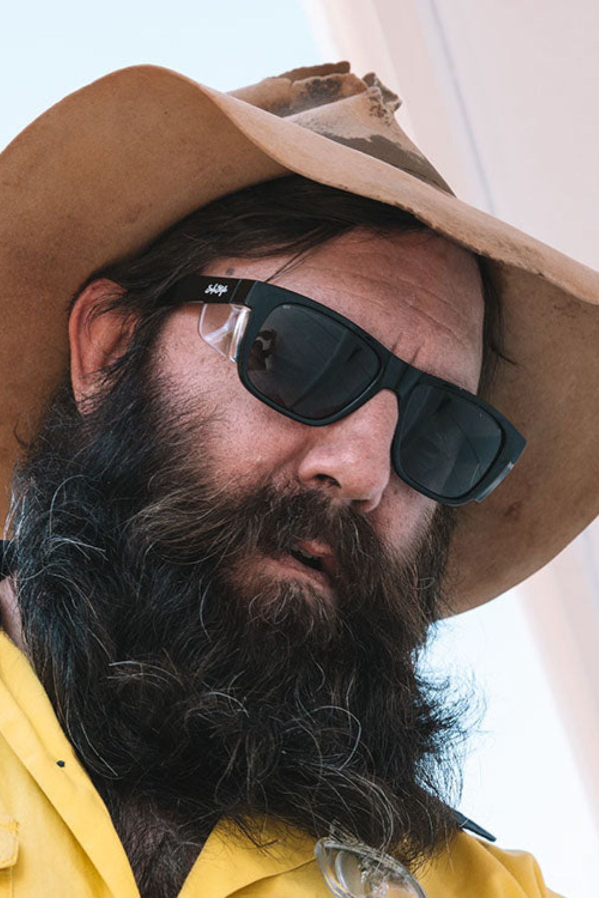 A man with a long, thick beard wears SafeStyle Fusions XL Black Frame Tinted Lens sunglasses, a wide-brimmed hat, and a yellow shirt while outdoors in bright light.